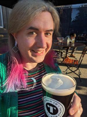 Non-binary person with hair of three colours, sitting outside in the sunshine holding a pint of beer and wearing a green cardigan