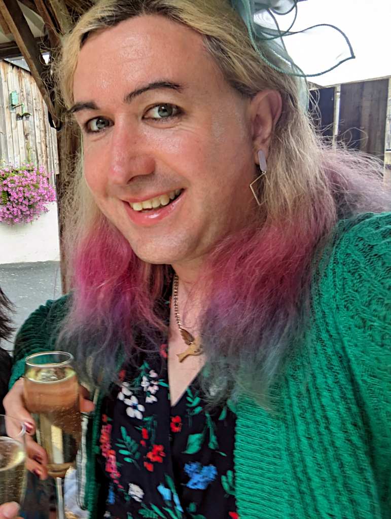 A picture a transfeminine person with brightly coloured hair at a wedding, wearing a dress and cardigan and with painted fingernails