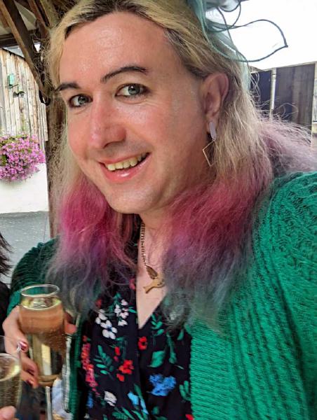A picture a transfeminine person with brightly coloured hair at a wedding, wearing a dress and cardigan and with painted fingernails
